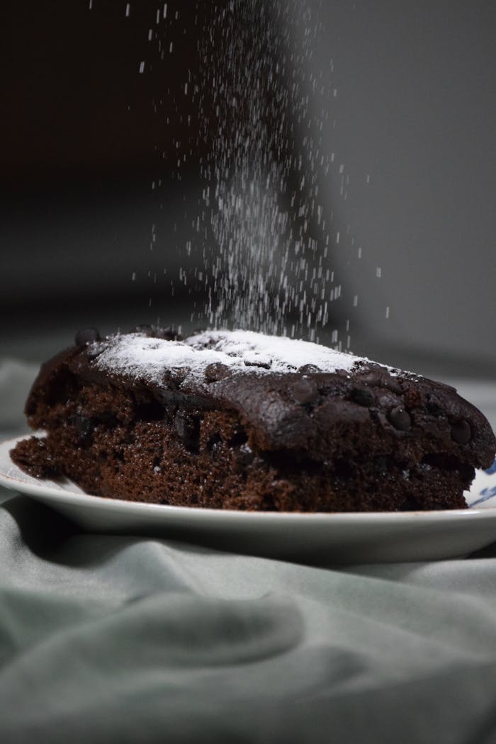 Close-up of chocolate cake slice being dusted with powdered sugar on plate.